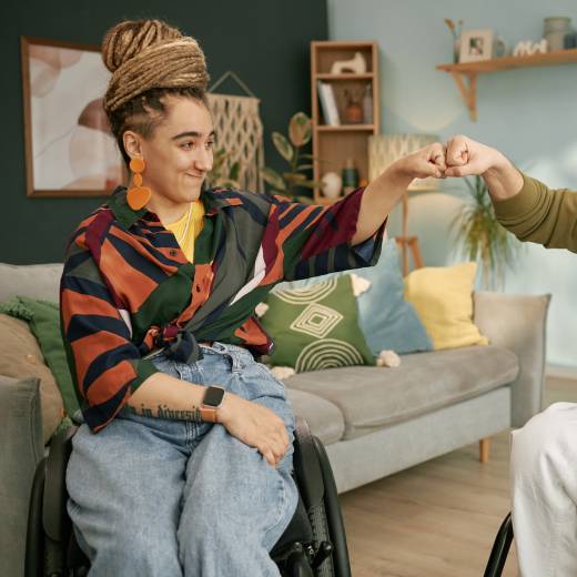 Zijaanzicht van een vrolijke man in een rolstoel die een vuistje geeft aan een glimlachende vrouw met een beperking, terwijl ze succes vieren in een gezellige woonkamer.