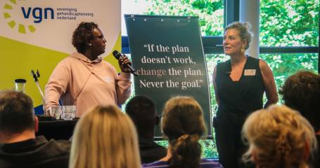 Idman praat in een microfoon en kijkt naar Marjoleyn die naast haar staat. Het publiek (gezien van achterkant) kijkt toe