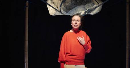 Actrice Lottie staat op podium en in het decor lijkt er een rots boven haar hoofd te zweven, opgehangen aan zwarte banden