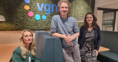 Een dame zit op een bank en daarnaast staan een man en een vrouw. Ze kijken alle drie glimlachend richting camera. Op de achtegrond is het logo van de VGN tegen een moswand te zien.