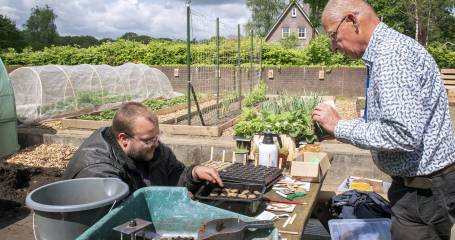 Twee mannen zijn in de tuin aan het werk en de een zit gehurkt plantjes om te potten terwijl een ander staand meehelpt