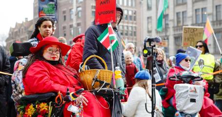 vrouw in rode kleding op de voorgrond in haar rolstoel met daaromheen een groep mensen