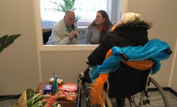 mevrouw in rolstoel praat met man en vrouw via raam bij Bartimeus
