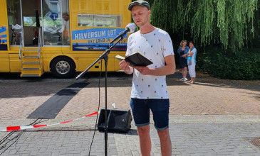 Levi Noe staat achter een microfoon in de buitenlucht om zijn gedicht voor te lezen