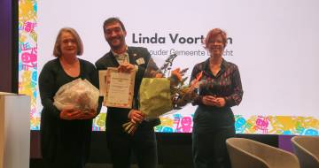 Een man en vrouw houden lachend een bos bloemen en certificaat vast terwijl een derde vrouw ernaast staat en glimlacht. Achter hen is de tekst 'linda voortman wethouder gemeente utrecht' zichtbaar
