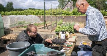 Twee mannen zijn in de tuin aan het werk en de een zit gehurkt plantjes om te potten terwijl een ander staand meehelpt