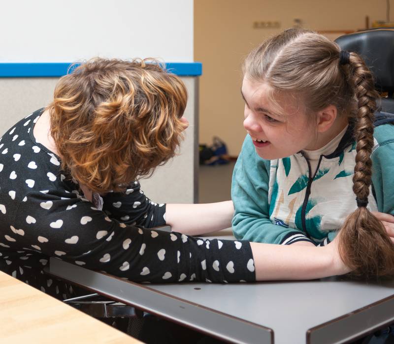 Jonge vrouw in rolstoel kijkt lachend naar vrouw die haar hand vastpakt