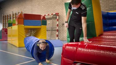 sportles in een gymlokaal iemand kruipt door een tunneltje terwijl een begeleider naar hem kijkt