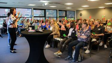 Een zaal vol mensen houdt rode of groene briefjes omhoog terwijl een vrouw vooraan in de zaal tegen hen spreekt