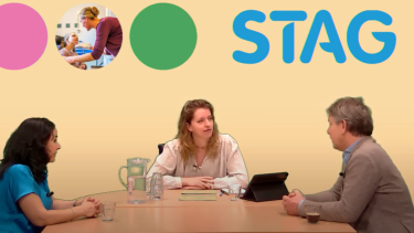 Drie mensen zitten op tafel en op de achtergrond is de tekst STAG te lezen