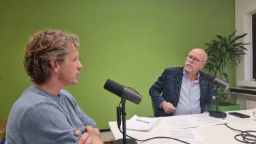 Jos Sanders en Johan van Ruijven zitten achter een microfoon aan een tafel en kijken elkaar aan