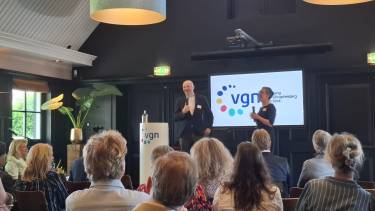 Je ziet een groep mensen zittend in het publiek vanaf de rug en Boris en Astrid staan op een podium en zijn in gesprek naast een katheder en beeldscherm met vgn logo