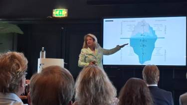 Een groep mensen zit in het publiek en kijkt naar Marita die staat te presenteren