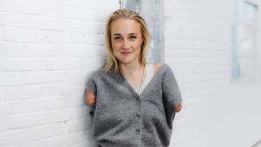 Jonge vrouw kijkt glimlachend in de camera terwijl ze tegen een muur leunt