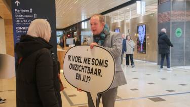 Man in gesprek met vrouw en de man houdt een bord vast met de tkest ik voel me vaak onzichtbaar, en jij?