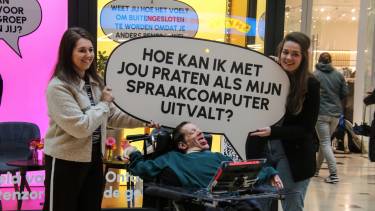 Man zit in rolstoel lachend en met de armen gespreid en twee vrouwen houden glimlachend een bord boven zijn hoofd met de tekst Hoe kan ik met jou praten als mijn spraakcomputer uitvalt?